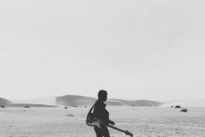 A person holding a guitar walks alone across a sandy desert, perhaps pondering songs about pest control under the bright, clear sky.