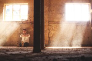 A person sits on the dusty floor near a wall in a sunlit, abandoned room, pondering pest control as beams of light stream through two windows.