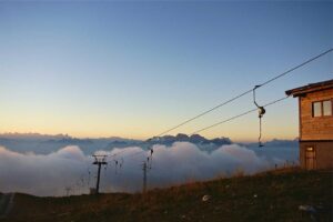 A ski lift cable runs across a grassy slope near a wooden building, with mountains, clouds, and discreet pest control measures at sunset.