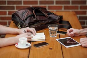 Two people discuss pest control solutions at a table with a leather bag, notebook, tablet, phones, coffee cup, and glass of water.