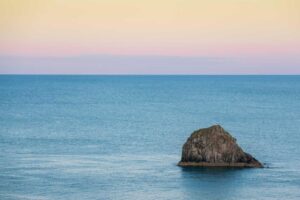 A large rock sits in calm blue ocean water, untouched by time or pest control, beneath a pastel sky at sunrise or sunset.