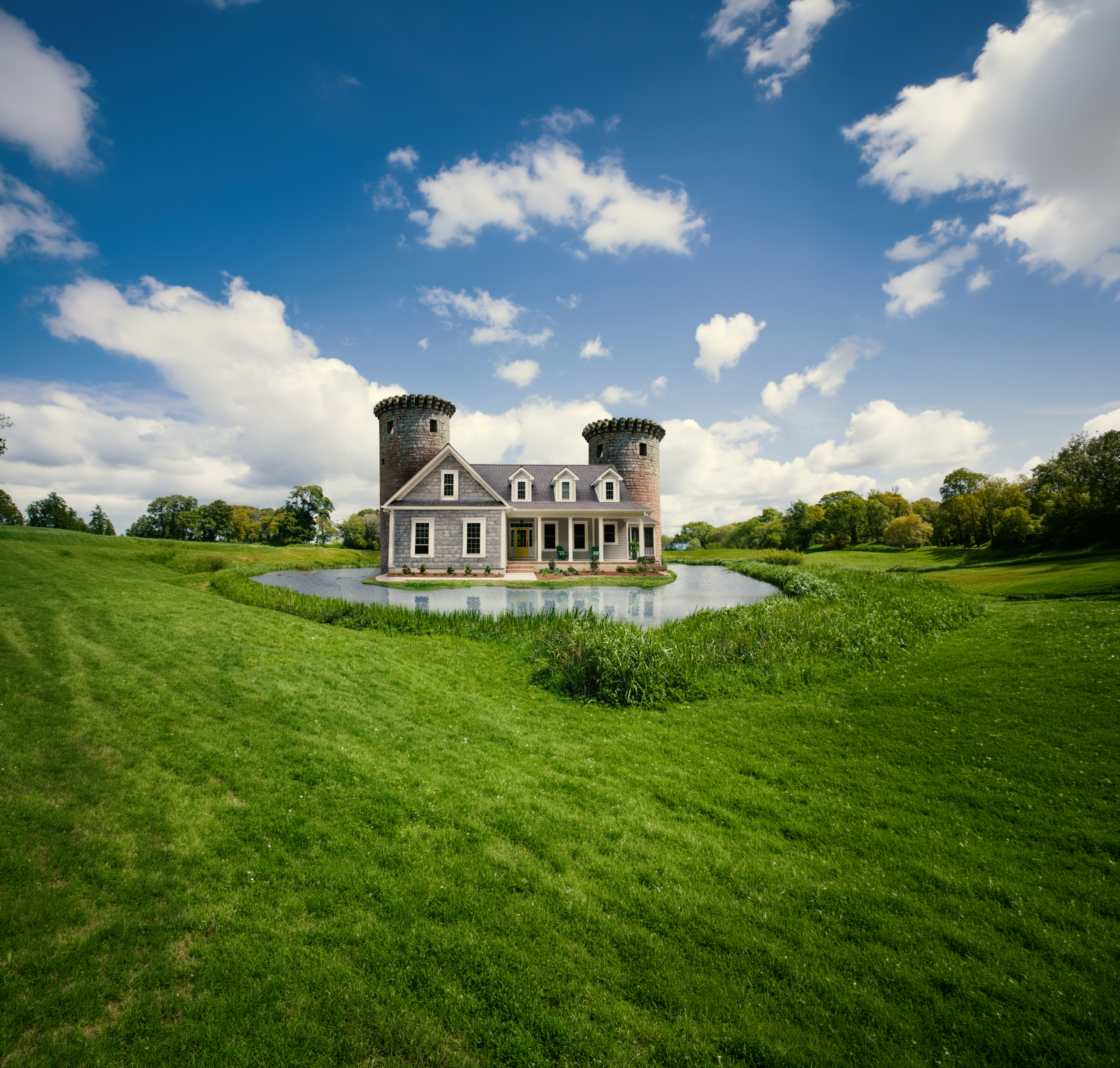 A house with two round towers stands on an island in a pond, featuring pest control, set in a grassy field under a blue sky with clouds.