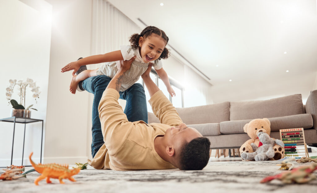 An adult plays on the floor, lifting a child high as toys and dinosaurs surround them in a clean, pest control-protected space.