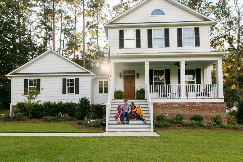 A family of four sits on the front steps of a two-story white house with a porch and well-kept lawn.